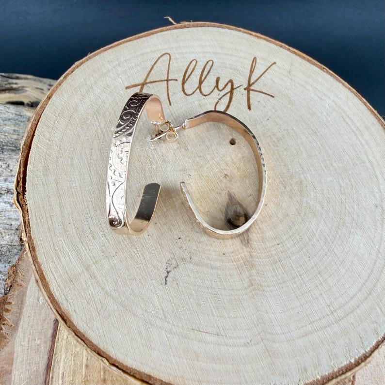 May include: A pair of gold-colored hoop earrings. One earring features an etched floral design, while the other has a textured surface. The earrings are displayed on a wooden surface with the name "Allyk" written on it.