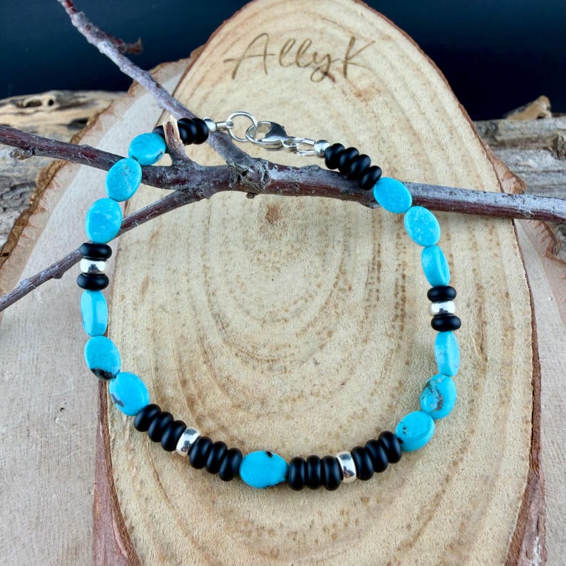 May include: A turquoise and black beaded bracelet with a silver clasp. The bracelet features oval turquoise stones interspersed with black beads and silver accents. The bracelet is displayed on a wooden surface.