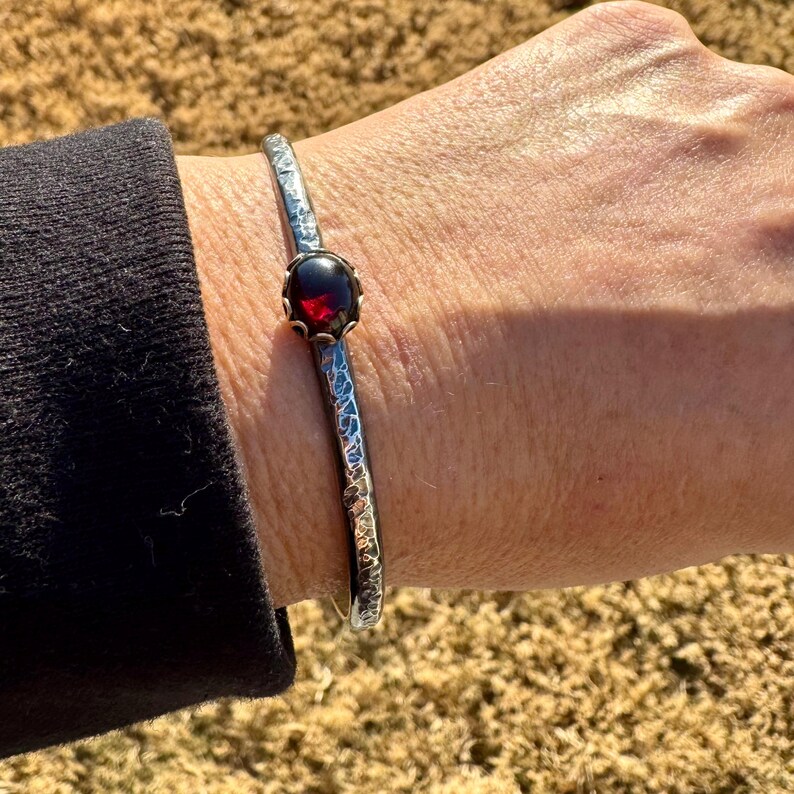 Puede incluir: Un brazalete plateado con textura martillada y una piedra preciosa de color rojo oscuro. La piedra preciosa est&aacute; engastada en un bisel decorativo. El brazalete se lleva en la mu&ntilde;eca.
