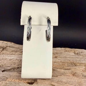May include: A pair of silver-toned hoop earrings with a textured, leaf-like pattern. The earrings are displayed on a white stand against a dark background, showcasing their intricate design and metallic sheen. The hoops are approximately 1 inch in diameter.