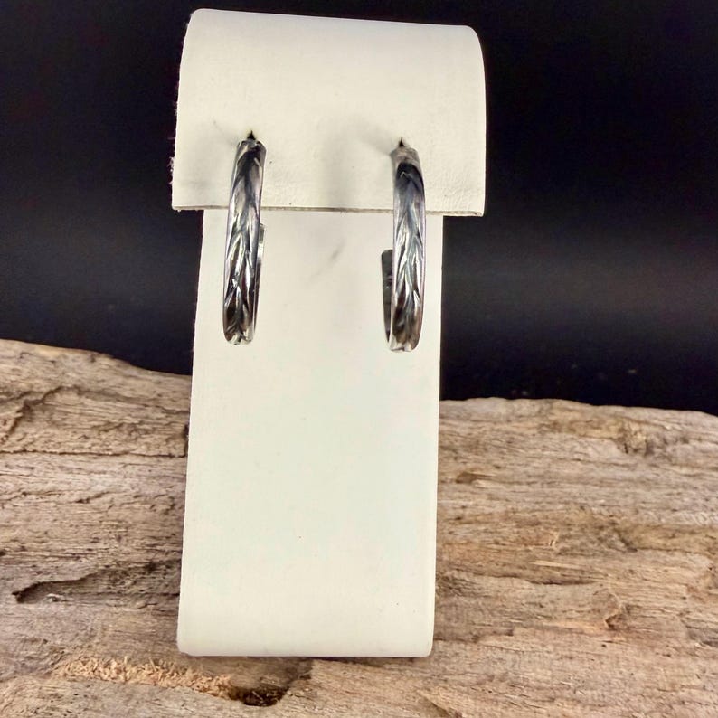 May include: A pair of silver-toned hoop earrings with a textured, leaf-like pattern. The earrings are displayed on a white stand against a dark background, showcasing their intricate design and metallic sheen. The hoops are approximately 1 inch in diameter.