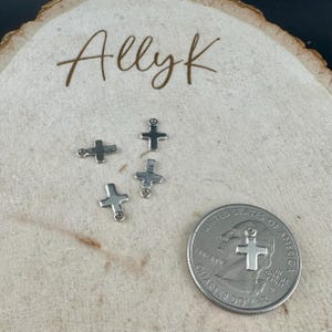 May include: Four small silver-colored cross charms with a quarter for size comparison. The charms are on a light-colored wooden surface with the handwritten text "Allyk" in brown script.