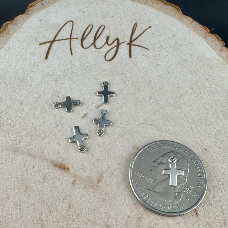 May include: Four small silver-colored cross charms with a quarter for size comparison. The charms are on a light-colored wooden surface with the handwritten text "Allyk" in brown script.