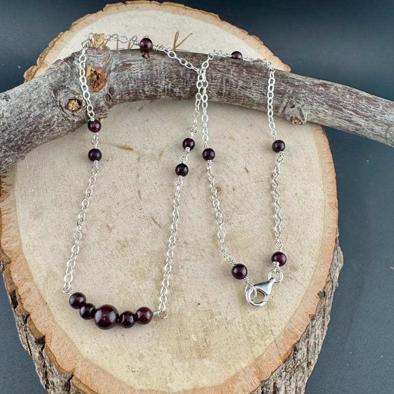 May include: A silver necklace with dark red garnet beads. The necklace features a delicate chain with several small beads and a cluster of larger beads at the center. A silver clasp secures the necklace.