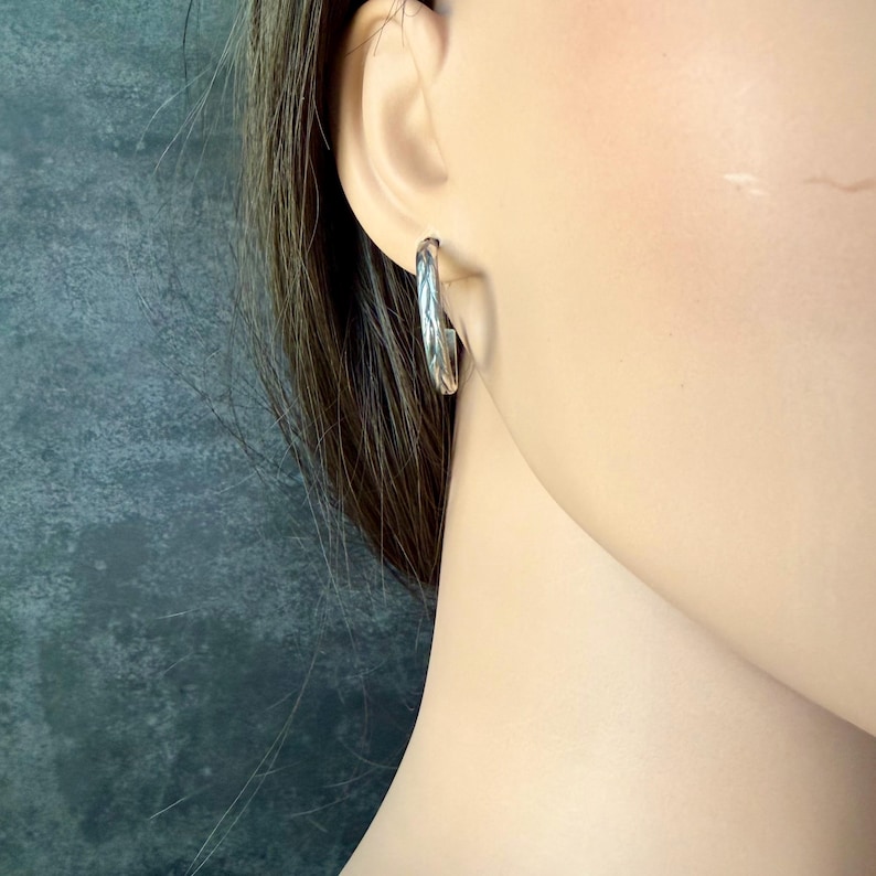 May include: A close-up of a silver hoop earring. The earring is a simple, polished design with a textured surface. The earring is worn on a person's ear, with the focus on the jewelry.
