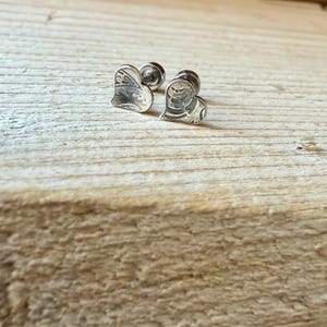 May include: A pair of silver-toned stud earrings shaped like hearts. Each heart features a textured, etched surface pattern and a small, cylindrical post backing attached to the center of the heart shape for secure placement.