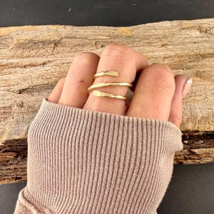 May include: A gold-colored ring with a spiral design. The ring wraps around the finger with three bands, each with a slightly textured surface. The ring is worn on a hand, with a neutral-colored sweater sleeve in the foreground.