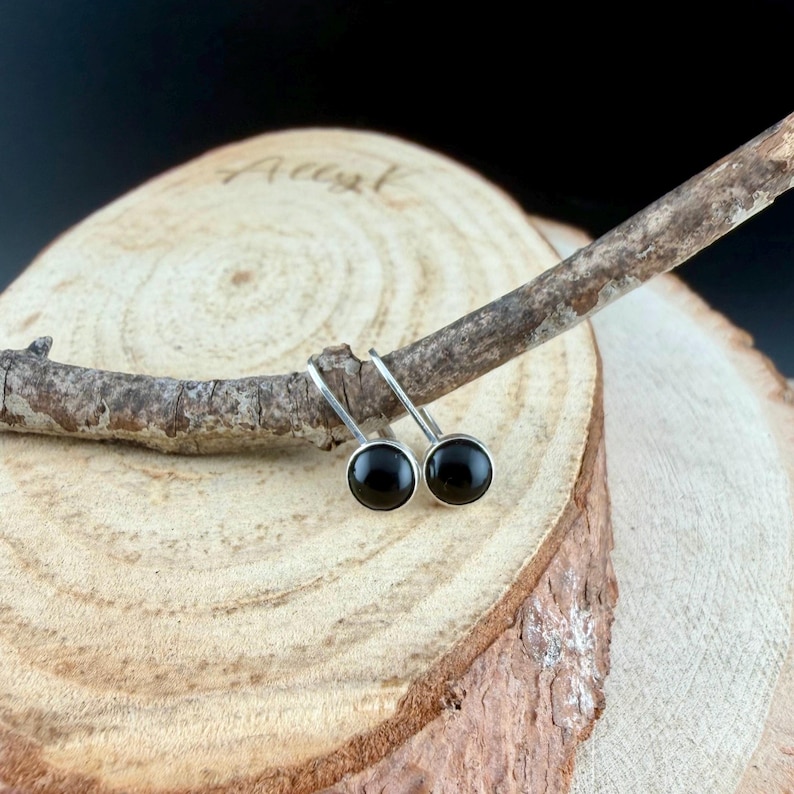 May include: A pair of small, round black earrings with silver-colored hooks. The earrings are displayed on a small, light brown wooden branch, set against a textured wooden background. The earrings have a simple, minimalist design.