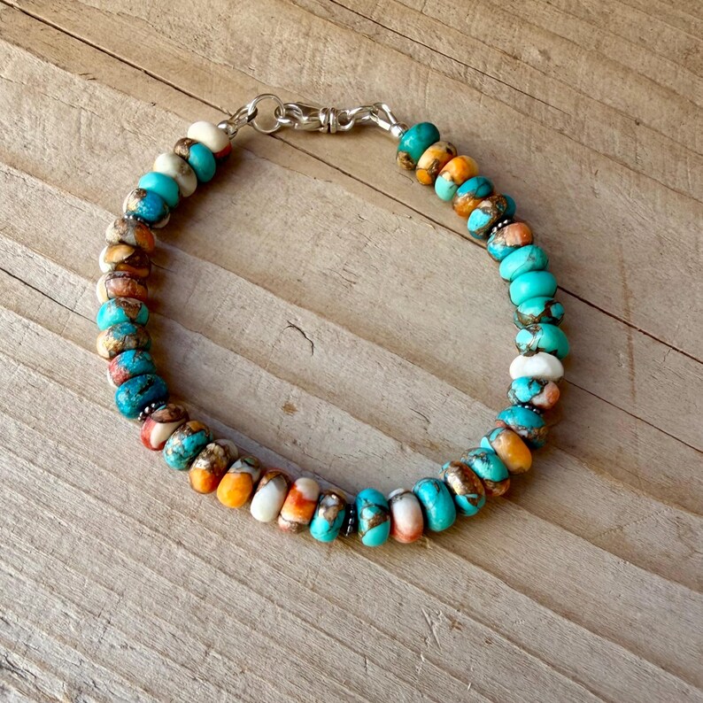 Pode incluir: Uma pulseira de contas com pedras redondas em forma de disco em tons vibrantes de turquesa, laranja, branco e castanho. As contas est&atilde;o enfiadas com pequenos espa&ccedil;adores de metal prateado e presas com um fecho de mosquet&atilde;o prateado.