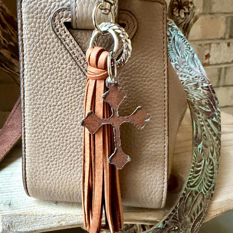 Op de afbeelding: Een beige leren kwastje sleutelhanger met een bruine kruisbedel met een verweerde metalen rand. Het kwastje is bevestigd aan een zilverkleurige metalen ring en karabijnhaak, hangend tegen een gestructureerde beige leren handtas aan.