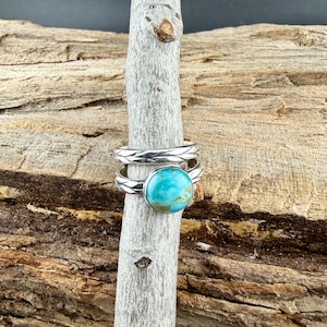 May include: Two silver rings, one with a turquoise stone and the other with a braided design. The rings are displayed on a wooden stick against a wood background. The turquoise stone is a vibrant blue-green color.