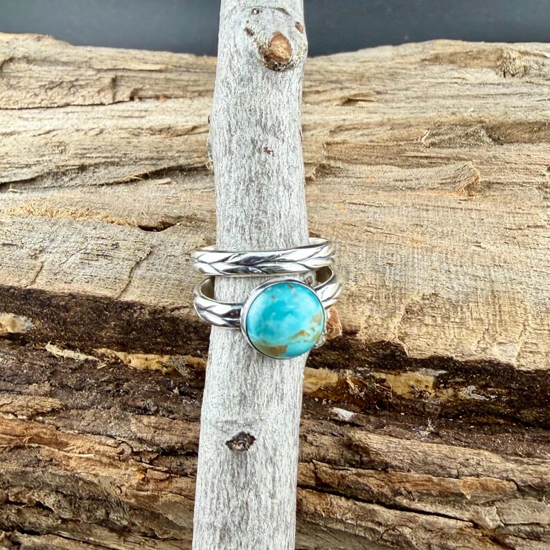 May include: Two silver rings, one with a turquoise stone and the other with a braided design. The rings are displayed on a wooden stick against a wood background. The turquoise stone is a vibrant blue-green color.