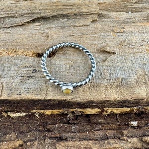 May include: A silver-colored ring with a twisted band design and a small, round, light yellow gemstone. The ring is set against a textured, brown wooden surface.