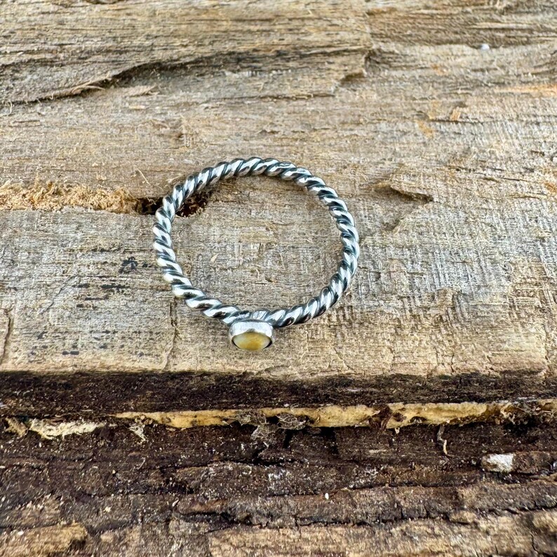 May include: A silver-colored ring with a twisted band design and a small, round, light yellow gemstone. The ring is set against a textured, brown wooden surface.