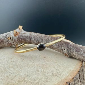 May include: A gold-toned bangle bracelet with a black oval stone set in the center. The bracelet is displayed on a wooden surface with a dark background. The simple design of the bracelet makes it a versatile accessory.
