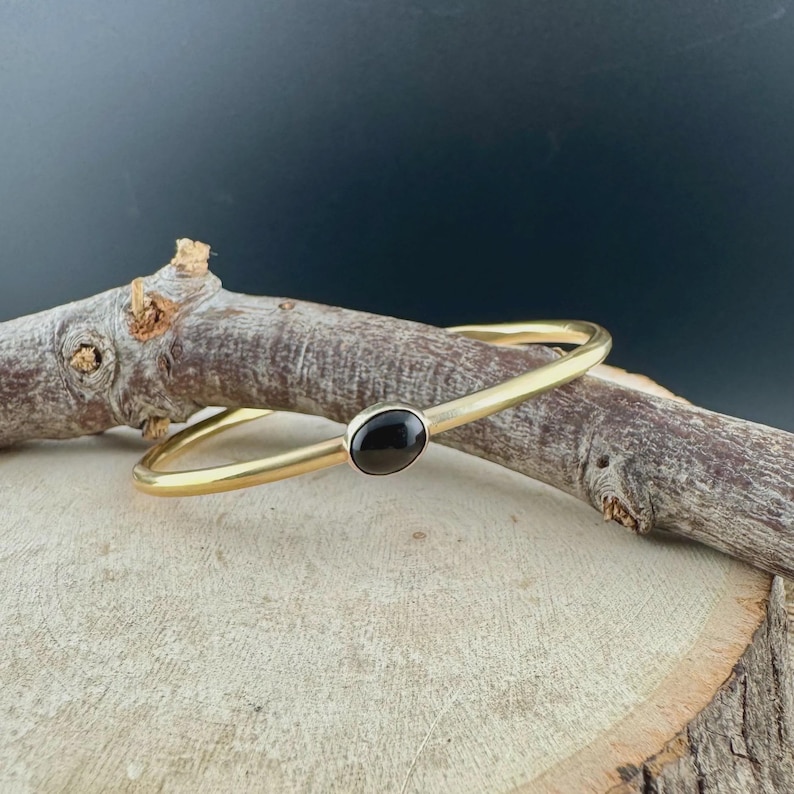 May include: A gold-toned bangle bracelet with a black oval stone set in the center. The bracelet is displayed on a wooden surface with a dark background. The simple design of the bracelet makes it a versatile accessory.