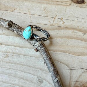 Puede incluir: Un anillo con una piedra de turquesa en forma de l&aacute;grima con vetas marrones, engastada en una banda de metal plateado con un dise&ntilde;o trenzado y retorcido. La piedra es de color azul claro con marcas naturales y la banda tiene un acabado oxidado.