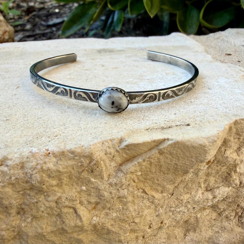May include: A silver cuff bracelet featuring an oval, speckled white gemstone. The bracelet has an open design and is decorated with an etched pattern. The gemstone is set in a decorative bezel.