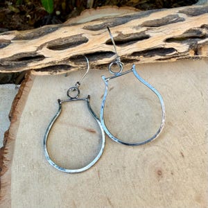 May include: A pair of silver-toned teardrop earrings with a hammered texture. The earrings have a simple, elegant design with a hook closure. The earrings are displayed on a wooden surface.