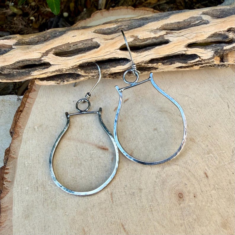 May include: A pair of silver-toned teardrop earrings with a hammered texture. The earrings have a simple, elegant design with a hook closure. The earrings are displayed on a wooden surface.