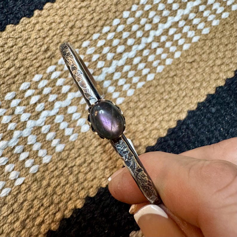 May include: A silver-toned bangle bracelet with a central oval-shaped, purple gemstone. The bracelet has an etched design along the sides. The background is a patterned fabric in shades of brown, black, and white.