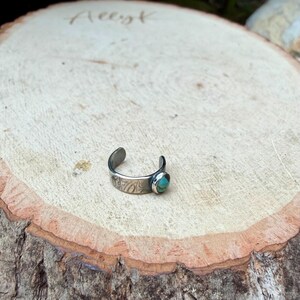K&ouml;nnte beinhalten: Ein verstellbarer Zehenring in Silber mit einem ovalen T&uuml;rkisstein. Das Band hat ein ge&auml;tztes Design. Der Ring wird auf einer hellbraunen Holzoberfl&auml;che gezeigt.