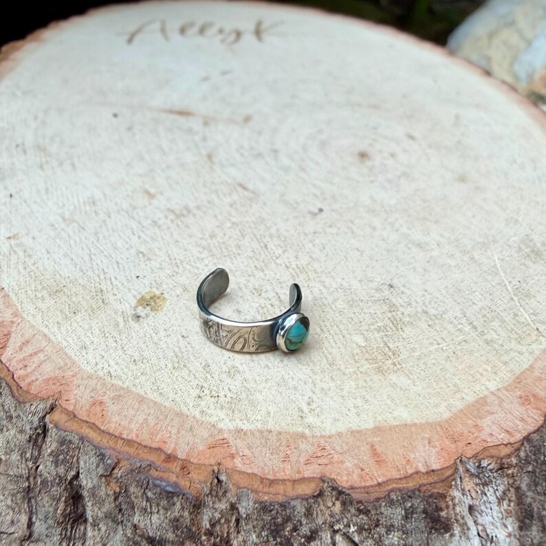 K&ouml;nnte beinhalten: Ein verstellbarer Zehenring in Silber mit einem ovalen T&uuml;rkisstein. Das Band hat ein ge&auml;tztes Design. Der Ring wird auf einer hellbraunen Holzoberfl&auml;che gezeigt.
