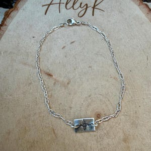 May include: A silver-colored chain bracelet with a rectangular pendant. The pendant features a cross design. The bracelet is displayed on a wooden surface. The word "Allyk" is written in cursive in the top left corner.