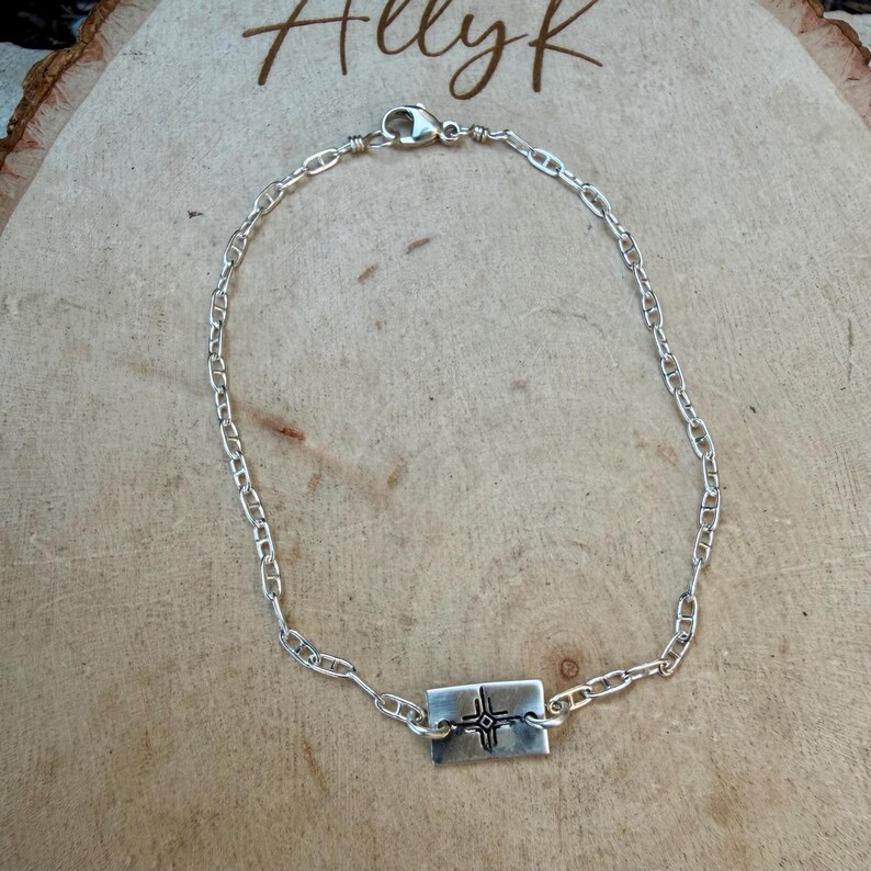 May include: A silver-colored chain bracelet with a rectangular pendant. The pendant features a cross design. The bracelet is displayed on a wooden surface. The word "Allyk" is written in cursive in the top left corner.