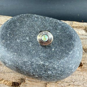 May include: A silver ring featuring a turquoise stone at its centre. The ring is crafted from a bullet casing, displaying the markings "40 S&W" and "W L N". The ring is placed on a grey stone.