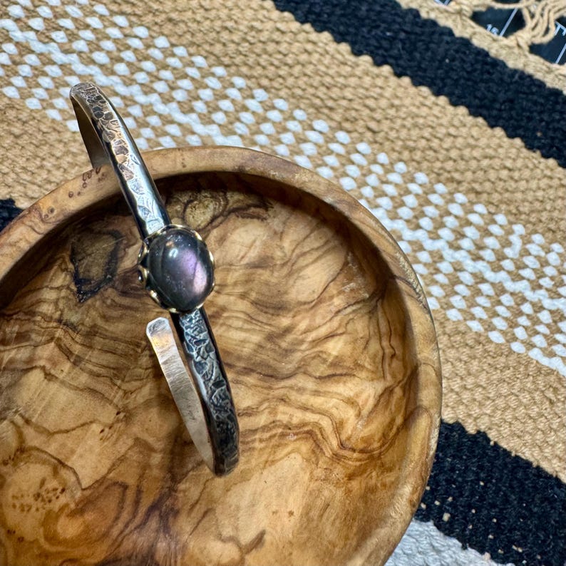 May include: Silver-toned cuff bracelet with a textured design and a large, oval, purple gemstone. The bracelet is open-ended and rests on a wooden bowl with a natural wood grain pattern. The background features a woven textile.