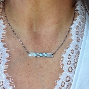 May include: A silver-toned necklace featuring a rectangular bar pendant with an intricate floral-like etched pattern. The bar is set with three small, smooth, light blue oval stones arranged horizontally along the center of the metal piece.
