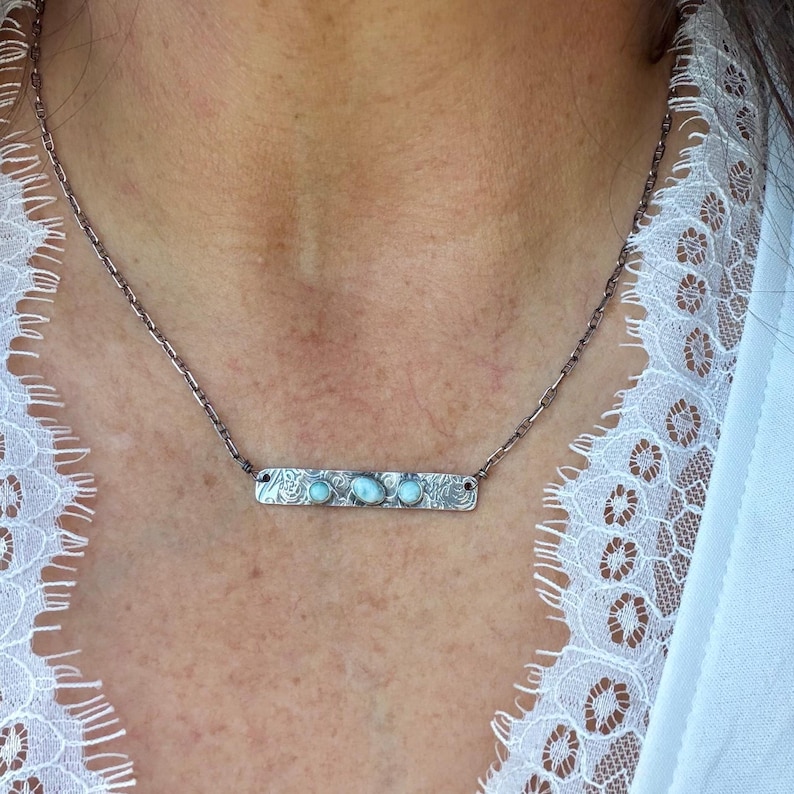 May include: A silver-toned necklace featuring a rectangular bar pendant with an intricate floral-like etched pattern. The bar is set with three small, smooth, light blue oval stones arranged horizontally along the center of the metal piece.