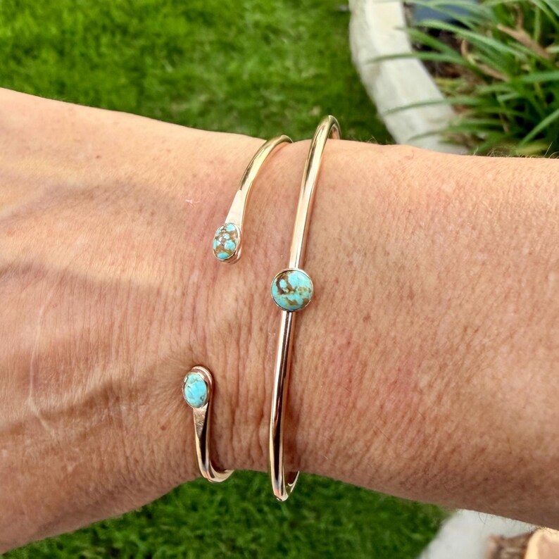 May include: Three gold-colored, open cuff bracelets with turquoise-colored stone accents. The bracelets are worn on a wrist, with the stones varying in size and placement. The background is blurred, with green grass visible.