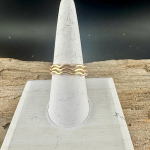 May include: A gold-colored ring with a wavy design. The ring is displayed on a white, cone-shaped stand. The ring's design features three stacked, undulating bands, creating a wave-like pattern. The background is dark.