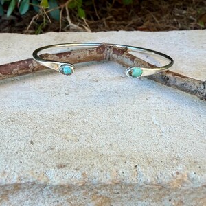 May include: A silver cuff bracelet featuring two small, oval-shaped turquoise stones set at each open end. The metal band has a smooth, polished finish, and the two light blue, speckled turquoise cabochons are securely mounted in silver settings.