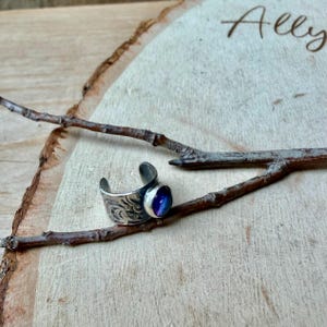 May include: A silver adjustable ring with a blue and purple stone. The ring has an engraved floral design on the band. The ring is displayed on a wooden surface with a branch.