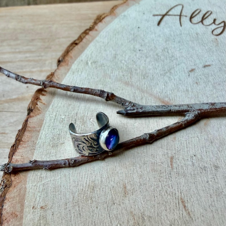 May include: A silver adjustable ring with a blue and purple stone. The ring has an engraved floral design on the band. The ring is displayed on a wooden surface with a branch.