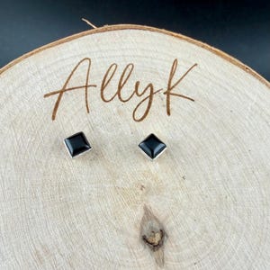 Puede incluir: Un par de pendientes de bot&oacute;n cuadrados negros con bordes plateados. Los pendientes se exhiben sobre una superficie de madera clara con el texto manuscrito "Allyk". El fondo es negro.