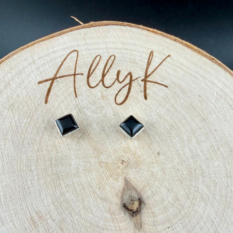 Puede incluir: Un par de pendientes de bot&oacute;n cuadrados negros con bordes plateados. Los pendientes se exhiben sobre una superficie de madera clara con el texto manuscrito "Allyk". El fondo es negro.