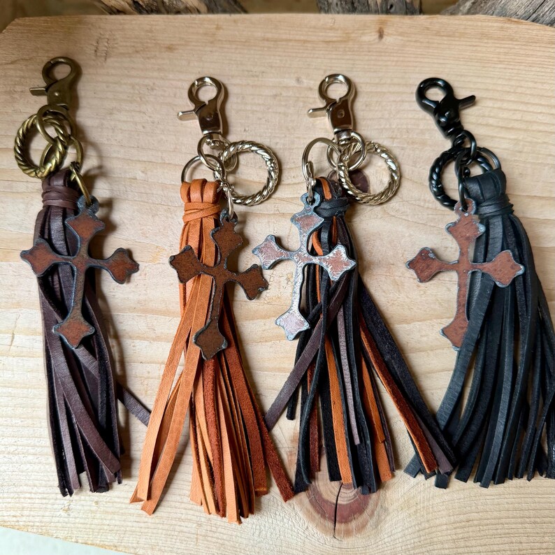 Op de afbeelding: Vier leren kwast-sleutelhangers met metalen kruisbedels. Elke hanger heeft een karabijnsluiting en een gedraaide ring. De kwasten zijn donkerbruin, lichtbruin, een mix van beide, en zwart, voorzien van kruisjes met een verweerde afwerking.