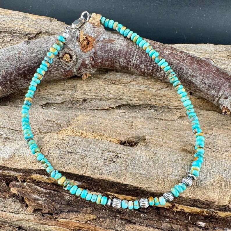 May include: An anklet featuring small, round turquoise beads strung together. The beads are a mix of blue and brown hues. Silver-colored metal beads and a clasp are also present. The anklet is displayed on a piece of wood.