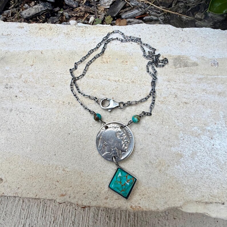 Op de afbeelding: Zilverkleurige ketting met een munt-hanger en een turquoise, ruitvormige steen. De ketting heeft een sluiting en kleine turquoise kralen. De munthanger heeft een afbeelding van een Indiaan.