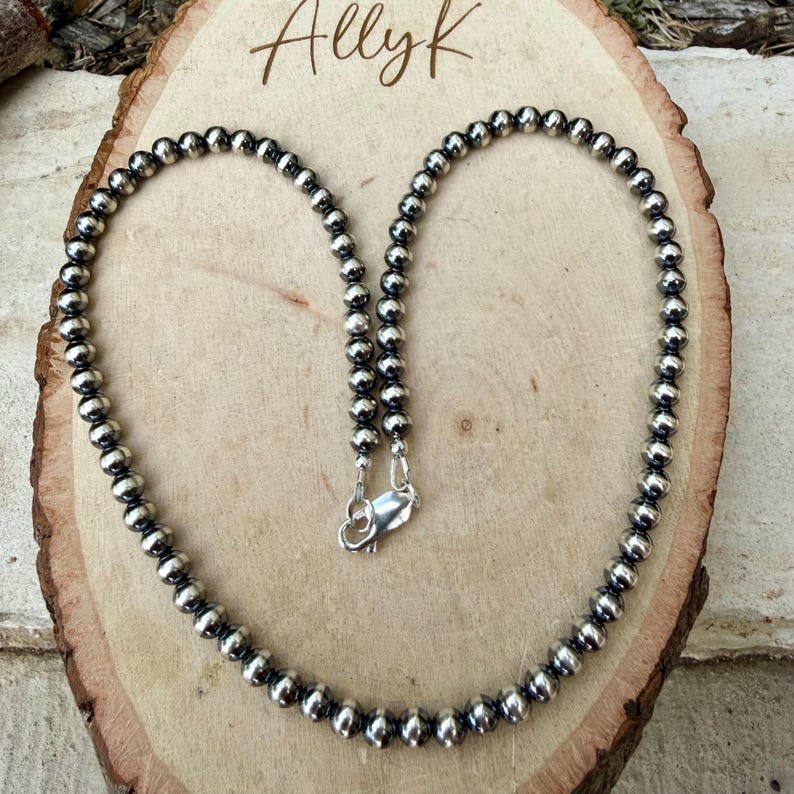 Pu&ograve; includere: Una collana di perline d'argento con chiusura a moschettone. La collana &egrave; composta da piccole perline rotonde d'argento. La collana &egrave; esposta su una superficie di legno. Il testo "Allyk" &egrave; scritto in corsivo nella parte superiore dell'immagine.