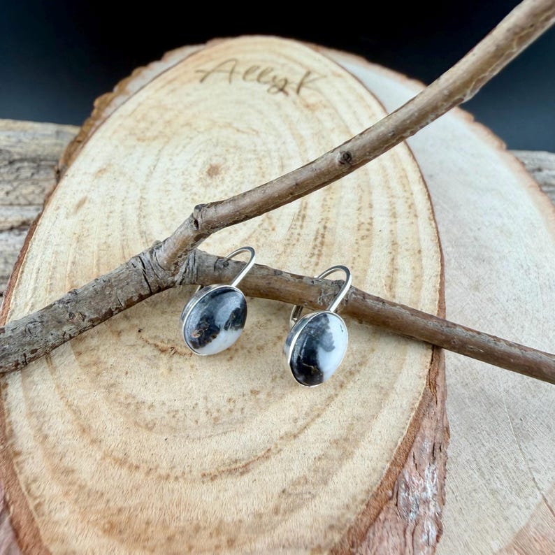 May include: A pair of oval-shaped earrings with a silver-colored metal setting. The earrings feature a black and white speckled stone. The earrings are displayed on a wooden surface with a small branch.