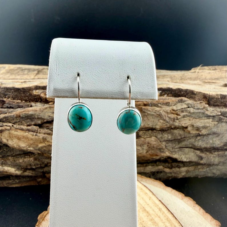 May include: A pair of turquoise earrings with a silver setting. The earrings are displayed on a white stand against a backdrop of wood. The turquoise stones are round and a vibrant teal color.