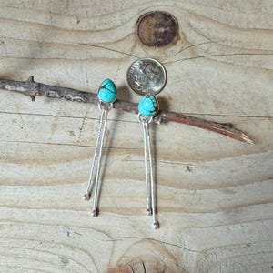 May include: A pair of dangle earrings featuring teardrop-shaped turquoise stones set in silver, with two long, thin, textured silver wires hanging from each stone, displayed next to a United States dime for scale reference.