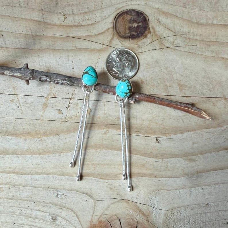 May include: A pair of dangle earrings featuring teardrop-shaped turquoise stones set in silver, with two long, thin, textured silver wires hanging from each stone, displayed next to a United States dime for scale reference.