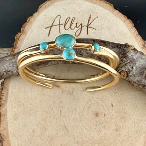 Op de afbeelding: Goudkleurige metalen manchetarmbanden met turkooizen stenen. De armbanden zijn gestapeld en hebben drie turkooizen stenen op de bovenste band. Het woord "Allyk" is in cursief op het houten oppervlak geschreven.