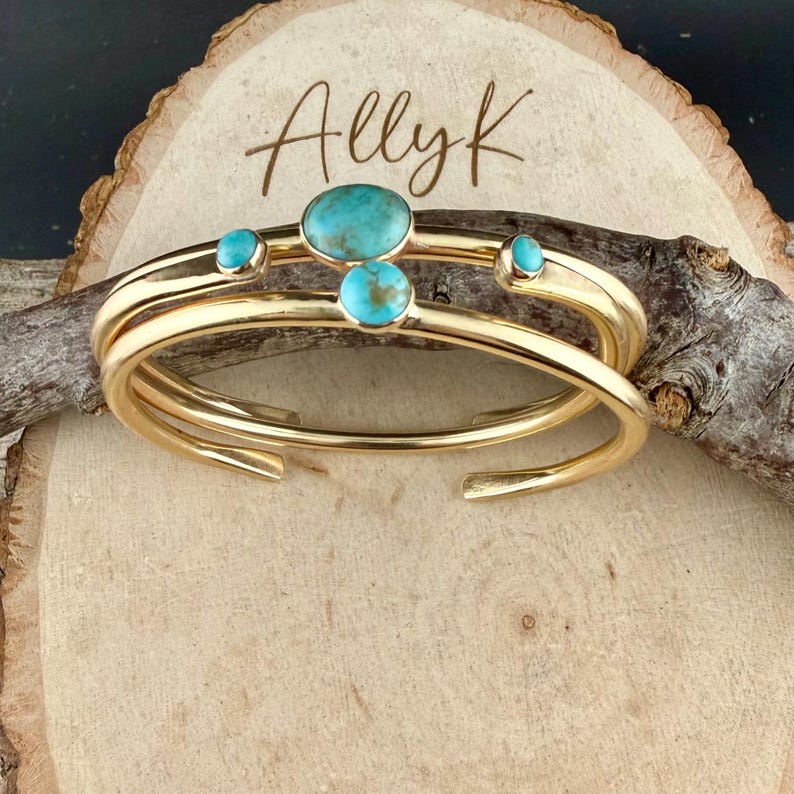 Op de afbeelding: Goudkleurige metalen manchetarmbanden met turkooizen stenen. De armbanden zijn gestapeld en hebben drie turkooizen stenen op de bovenste band. Het woord "Allyk" is in cursief op het houten oppervlak geschreven.